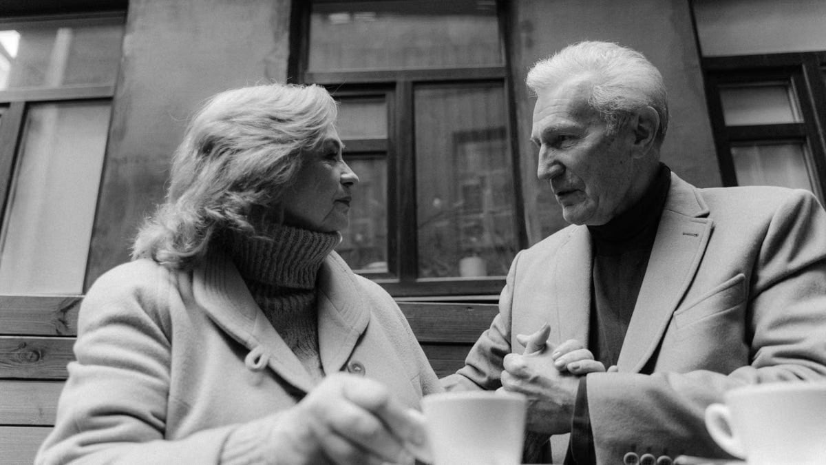 A black and white photo of a senior couple holding hands while having coffee outdoors.